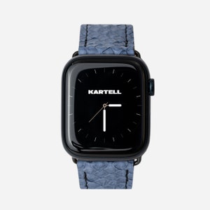Puede incluir: Un reloj inteligente negro con una correa de cuero azul texturizado. La esfera del reloj es negra con manecillas blancas y la palabra "KARTELL" en blanco. El reloj se muestra sobre un fondo blanco.