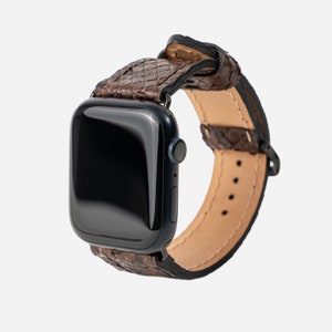 Puede incluir: Un reloj inteligente azul oscuro con una correa de reloj marrón con textura de piel de serpiente. La correa está hecha de un material marrón oscuro con un patrón de escamas. La correa está sujeta al reloj con herrajes metálicos negros. La esfera del reloj es negra.