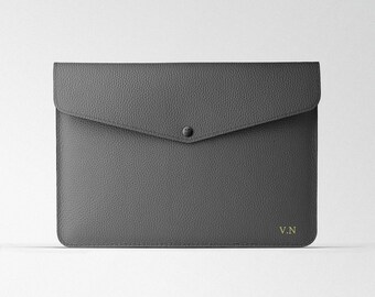 Puede incluir: Una funda para portátil de cuero gris con cierre de solapa y un solo botón a presión. La funda tiene las iniciales "V.N." grabadas en oro en la esquina inferior derecha.