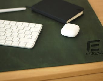May include: A green leather desk mat with a black leather notebook, a white wireless keyboard, a white wireless earbud case, and a black logo that says "Essence Creativity" with a stylized letter "E".