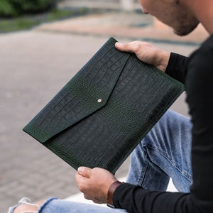 May include: A dark green leather envelope clutch with a crocodile embossed pattern. The clutch has a flap closure and a gold button.