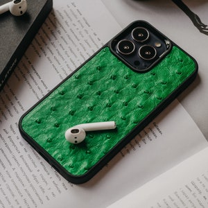 Pode incluir: Uma capa de telefone verde com um padrão texturizado que se assemelha a couro de avestruz. A capa tem uma borda preta e um recorte para as lentes da câmera do telefone.