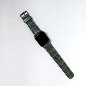 May include: A dark green leather watch band with a crocodile pattern. The band is attached to a black smartwatch with a black screen.