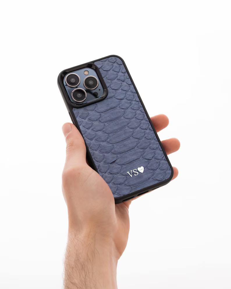 May include: A blue snakeskin phone case with a black border and a camera lens cutout. The case has the initials "VS" with a heart symbol in white.