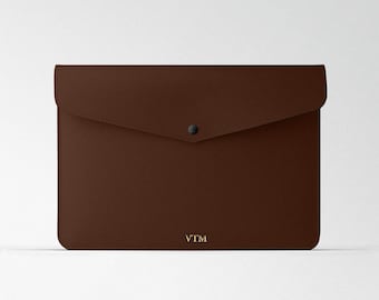 Puede incluir: Un clutch de cuero marrón en forma de sobre con cierre de solapa y un botón a presión único. El clutch tiene la palabra "VTM" grabada en oro en la parte delantera.