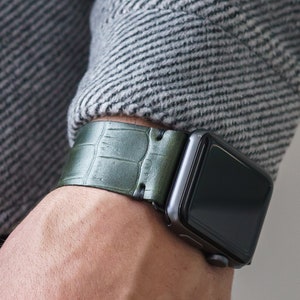 May include: A green leather watch band with a crocodile pattern. The band is attached to a black Apple Watch.