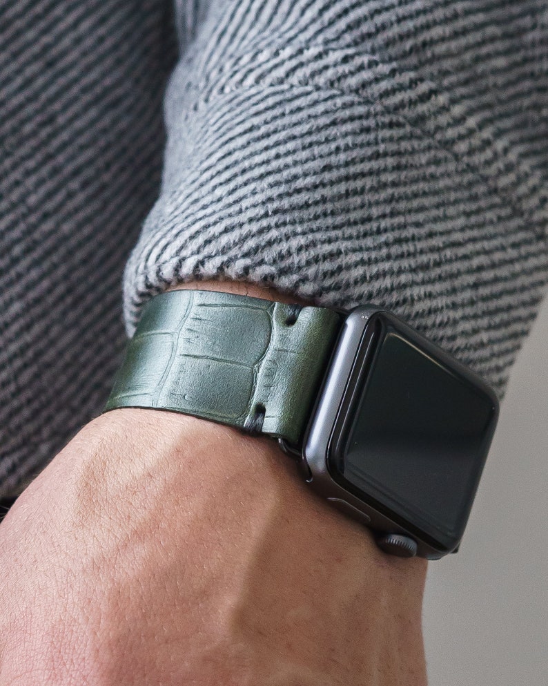 May include: A green leather watch band with a crocodile pattern. The band is attached to a black Apple Watch.