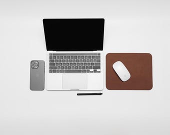 May include: A white laptop computer with a black keyboard and trackpad, a gray smartphone, a brown leather mousepad, and a white wireless mouse. The laptop is open and the screen is black.