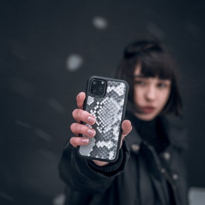 Może przedstawiać: Osoba trzymająca czarne etui na telefon z białym wzorem sk&oacute;ry węża.
