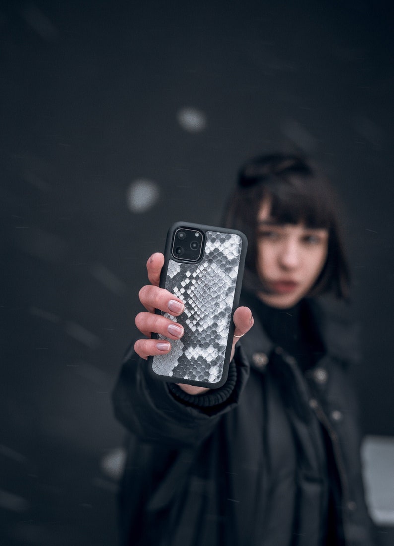 Może przedstawiać: Osoba trzymająca czarne etui na telefon z białym wzorem sk&oacute;ry węża.