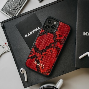 Może przedstawiać: Czerwona skórzana obudowa iPhone'a ze skóry węża z czarną obwódką i wytłoczonym białym napisem "ANGEL" na tylnej stronie.