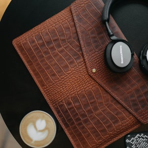 May include: A brown leather crocodile-embossed laptop sleeve with a flap closure and a gold button. Headphones with the text "Bowers & Wilkins" are on top of the sleeve.