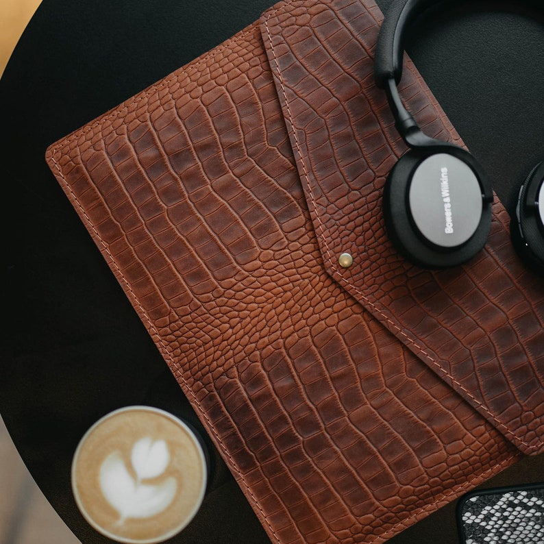 May include: A brown leather crocodile-embossed laptop sleeve with a flap closure and a gold button. Headphones with the text "Bowers & Wilkins" are on top of the sleeve.