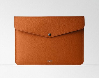 Puede incluir: Una funda para portátil de cuero marrón con cierre de solapa y un solo botón a presión. La funda tiene las iniciales "JMS" grabadas en la parte delantera.
