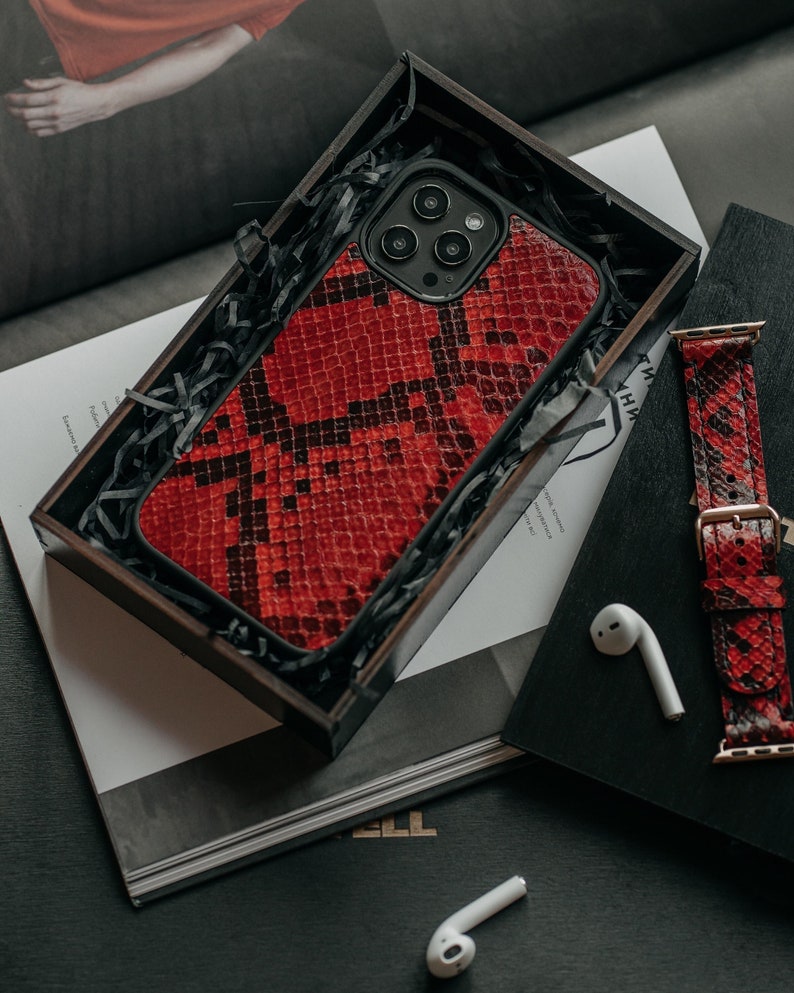 May include: A red snakeskin phone case with a black camera bump and a black border. The case is in a wooden box with shredded paper.