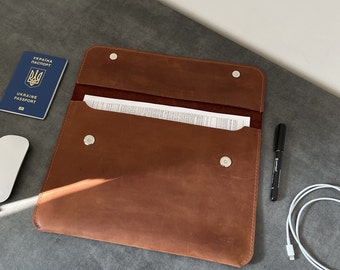 May include: A brown leather folder with a flap and a snap closure. The folder is open and has a white piece of paper inside. There is a black pen and a white cable on the table next to the folder.