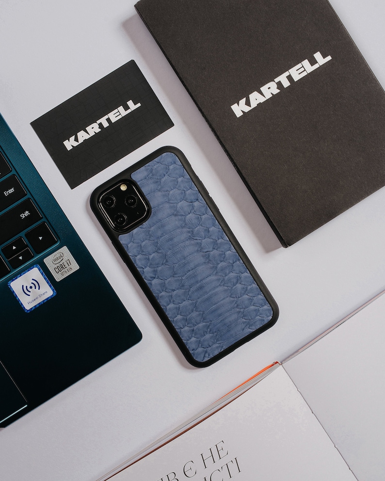 May include: A blue leather phone case with a snakeskin pattern and black trim. The case is on a white surface with a laptop, a business card, and a notebook.