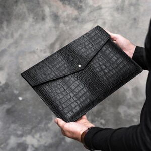 May include: Black leather envelope clutch with a crocodile embossed pattern. The clutch has a flap closure with a single button.