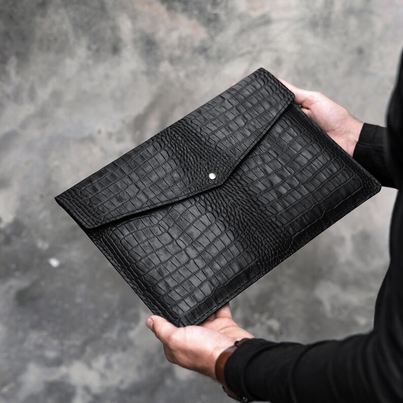 May include: Black leather envelope clutch with a crocodile embossed pattern. The clutch has a flap closure with a single button.