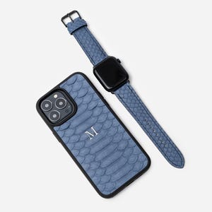 May include: A blue snakeskin iPhone case with a black border and the letter "M" monogrammed on the back. A matching blue snakeskin Apple Watch band with a black buckle is also pictured.