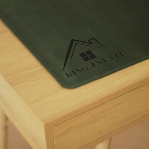 Puede incluir: Una alfombrilla de escritorio de cuero verde con el texto "KING ESTATE" en relieve en el centro. La alfombrilla está sobre un escritorio de madera clara con un cajón.