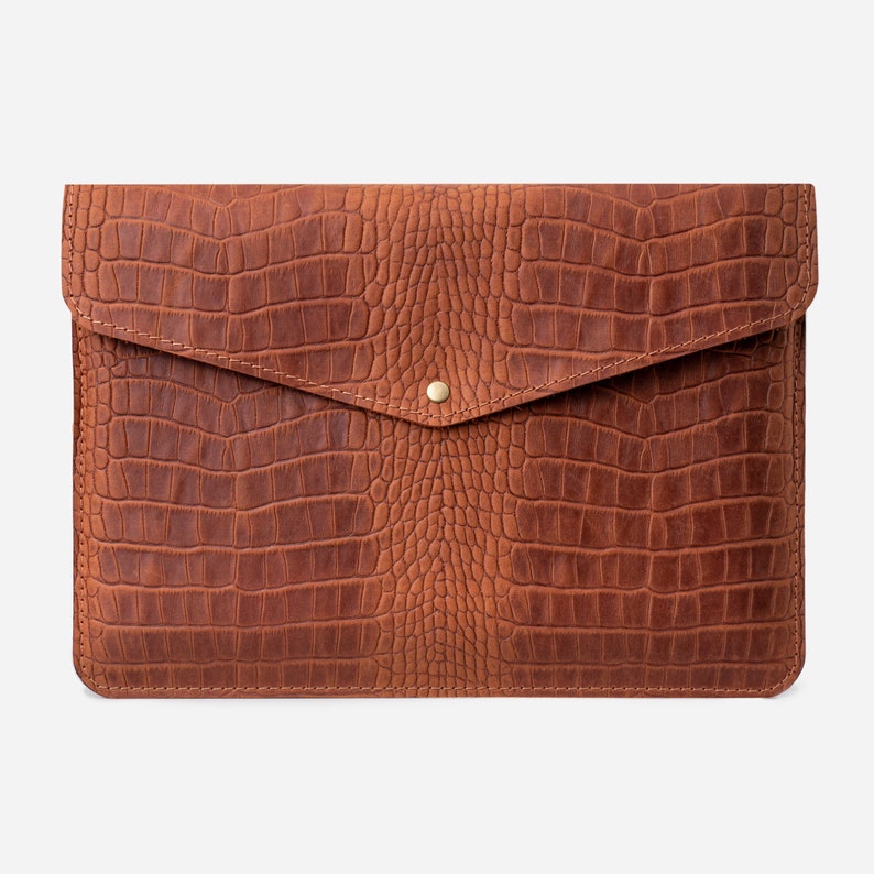 May include: A brown leather laptop sleeve with a crocodile embossed pattern. The sleeve has a flap closure with a brass button.