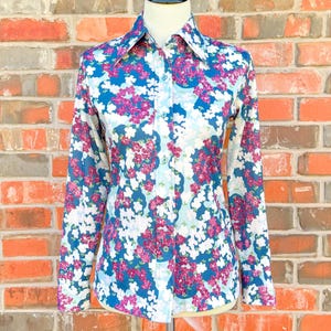 May include: A long-sleeve button-down shirt with a floral pattern. The shirt features a blue background with white and pink flowers. The shirt is displayed on a mannequin against a brick wall.