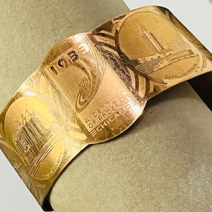 May include: A gold-toned metal cuff bracelet featuring engraved architectural illustrations and the text 1933 A CENTURY OF PROGRESS CHICAGO. The design includes detailed depictions of buildings and geometric patterns etched into the polished surface.