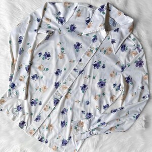 May include: White button-down shirt with a floral pattern. The shirt features a repeating design of purple and peach-colored roses with green leaves. The long-sleeved shirt has a classic collar and buttoned cuffs, and is made of a lightweight fabric.