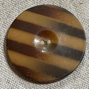 May include: A large, round button with a striped pattern in shades of brown and tan. The button has two holes for sewing and a slightly glossy finish. The button is a vintage style.