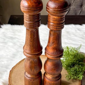 May include: Two wooden salt and pepper grinders with a rich, dark brown finish. The grinders have a classic design with a bulbous base, a ridged midsection, and a rounded top. They are displayed on a wooden slice, with a white fluffy surface in the background.