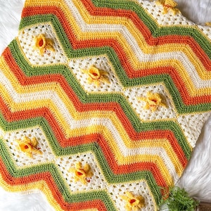 May include: A crocheted blanket with a chevron pattern in shades of yellow, orange, green, and white. The blanket has a textured surface with small flower-shaped details.