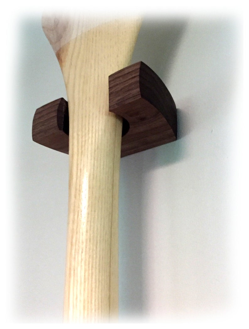 May include: A dark brown wooden wall mount bracket holding a light-colored wooden paddle. The bracket features a curved, semi-circular design that secures the shaft of the paddle against the wall, showcasing the natural wood grain textures.