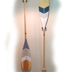 May include: Two wooden canoe paddles mounted vertically. The left paddle features a grey, white, and natural wood chevron pattern. The right paddle displays a blue, light grey, and white chevron design on the blade. Both have light wood shafts.