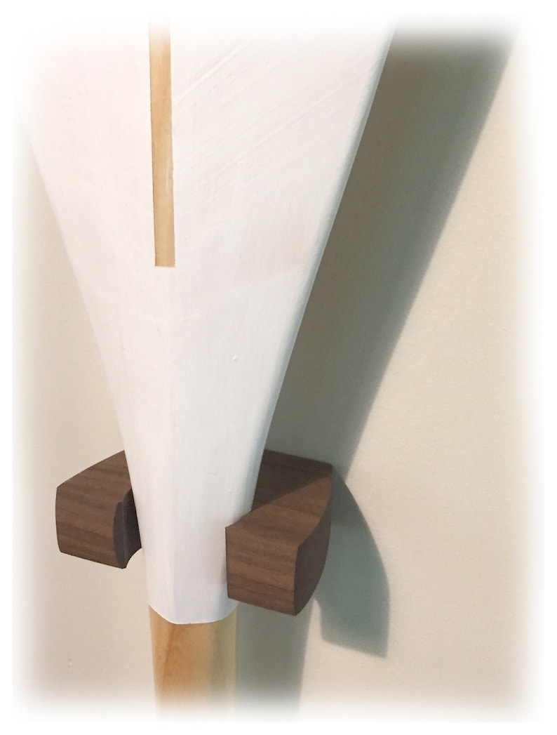 May include: A white painted wooden paddle blade with a visible natural wood vertical stripe, held in place by a dark brown wooden wall-mounted bracket. The bracket features two curved, block-like supports that cradle the lower section of the paddle.
