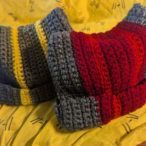 May include: Two handmade crochet beanies. One is gray with a yellow stripe, the other is gray with a red heart design. The beanies are displayed on a yellow surface.