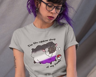 Op de afbeelding: Een persoon draagt een grijs T-shirt met een cartoon schaap en de tekst "Ik ben het regenboogschaap van mijn familie". Het schaap is wit met zwarte vlekken en heeft een paarse, witte en zwarte streep over zijn lichaam. De persoon heeft paars haar en draagt een bril.