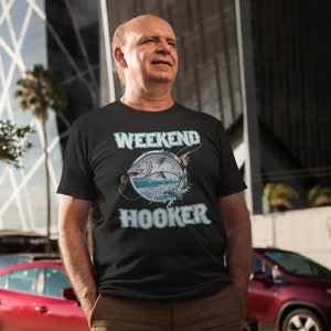 Puede incluir: Un hombre con una camiseta negra con un gráfico de un pez y el texto "Weekend Hooker".