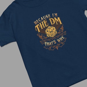 May include: A navy blue t-shirt with a gold design featuring a 20-sided die and the text "Because I'm the DM That's Why."