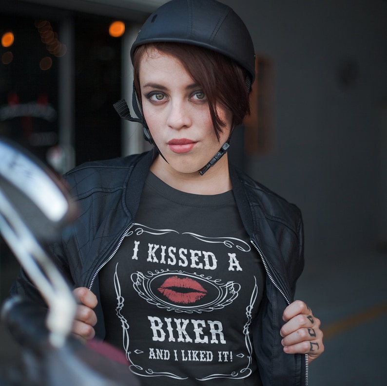 Peut inclure: Une femme portant une veste en cuir noire et un casque de moto noir avec un t-shirt blanc &agrave; imprim&eacute; graphique qui dit "J'ai embrass&eacute; un motard et j'ai aim&eacute; &ccedil;a !" avec un graphique de rouge &agrave; l&egrave;vres rouge.