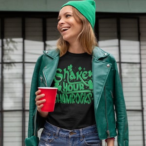 Op de afbeelding: Een vrouw draagt een groene beanie, een groen leren jack en een zwart t-shirt met de tekst "Shake Your Shamrocks" in groene letters. Ze houdt een rood plastic bekertje vast.
