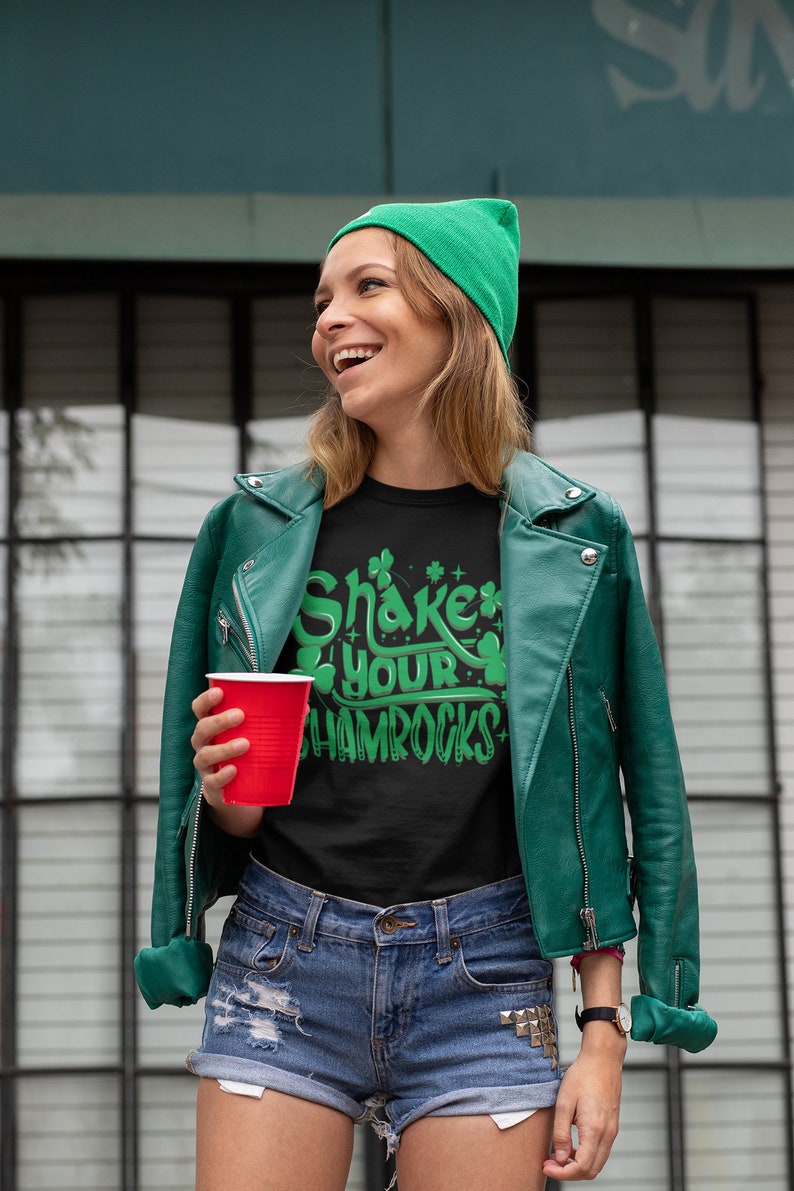 Op de afbeelding: Een vrouw draagt een groene beanie, een groen leren jack en een zwart t-shirt met de tekst "Shake Your Shamrocks" in groene letters. Ze houdt een rood plastic bekertje vast.