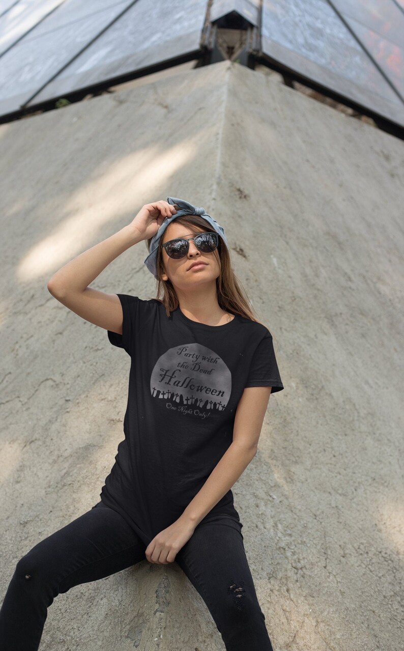 May include: A woman wearing a black t-shirt with a white graphic that says "Party with the Dead Halloween One Night Only!" She is wearing black pants and sunglasses.