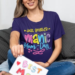 May include: A woman wearing a purple t-shirt with the text "Just another manic mom-day" in colorful letters. She is holding a white card with the text "Best Mom" in colorful letters.