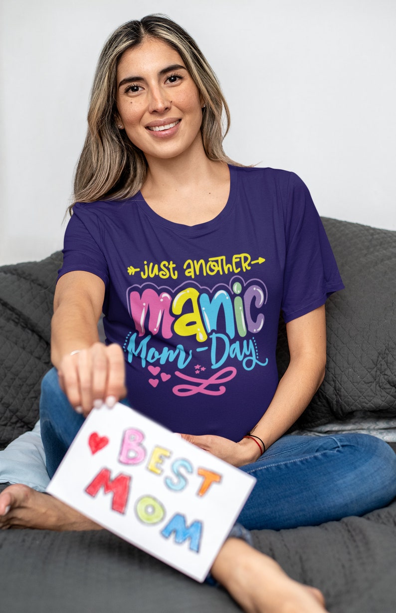 May include: A woman wearing a purple t-shirt with the text "Just another manic mom-day" in colorful letters. She is holding a white card with the text "Best Mom" in colorful letters.