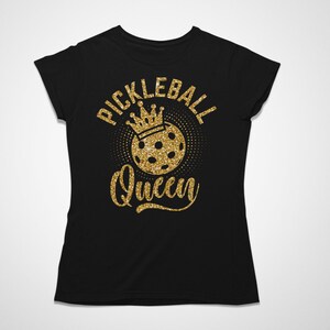May include: Black t-shirt with a gold glitter design. The design features the words "Pickleball Queen" in a script font, with a crown above a pickleball. The shirt is a crew neck style.