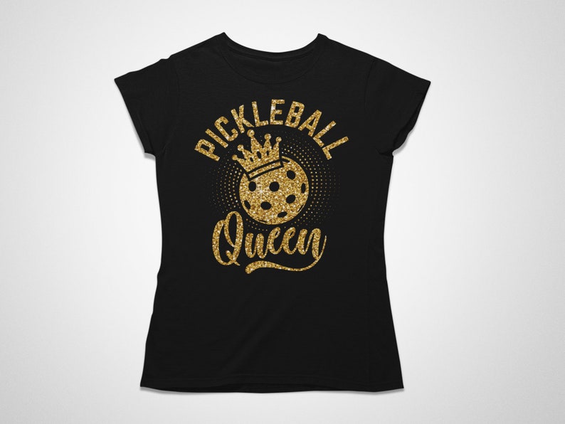 May include: Black t-shirt with a gold glitter design. The design features the words "Pickleball Queen" in a script font, with a crown above a pickleball. The shirt is a crew neck style.