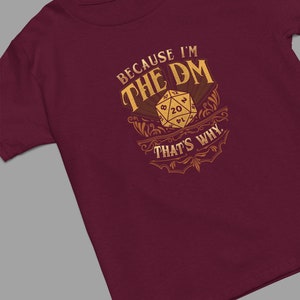 May include: Burgundy t-shirt with a gold design featuring a 20-sided die and the text "Because I'm the DM That's Why."