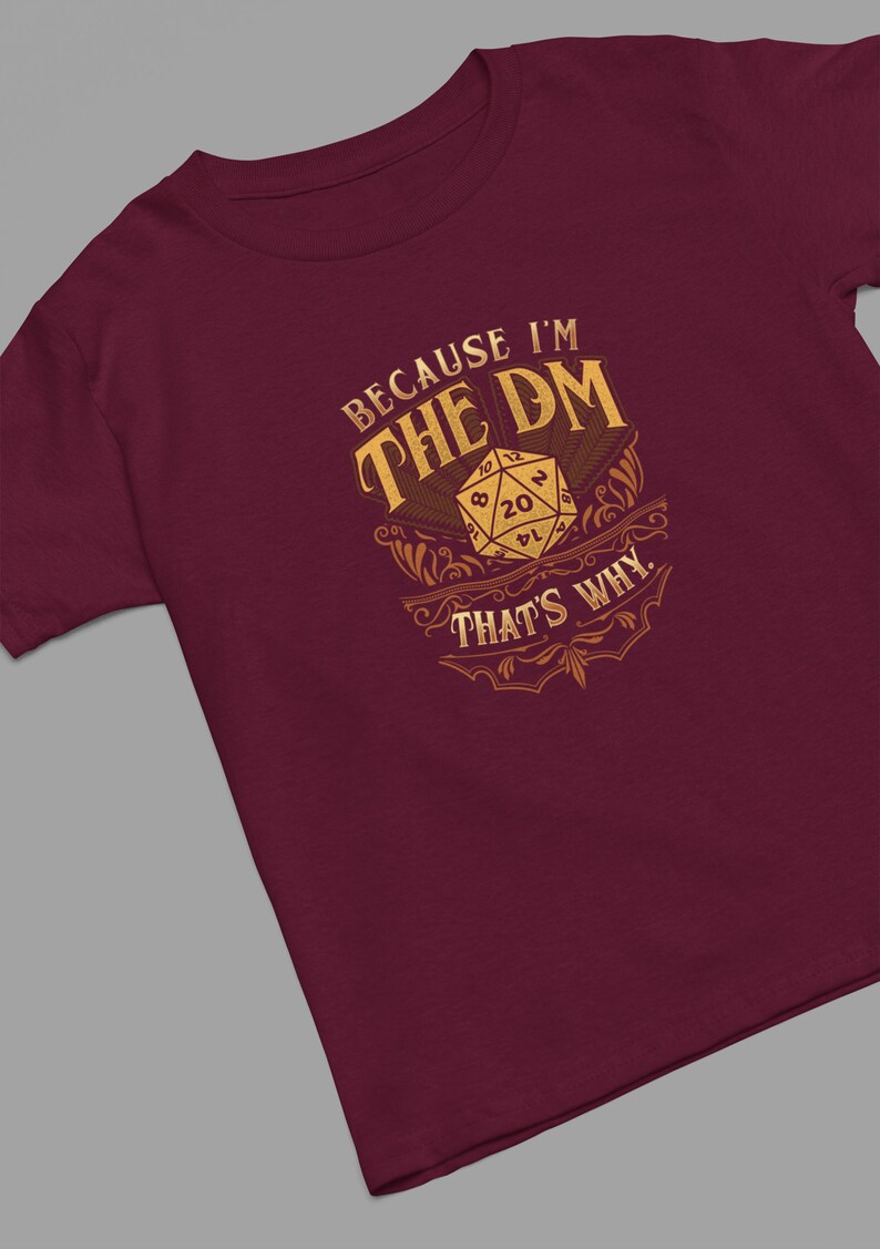 May include: Burgundy t-shirt with a gold design featuring a 20-sided die and the text "Because I'm the DM That's Why."