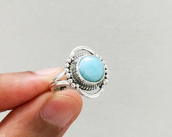 May include: A silver ring with a light blue gemstone set in a circular bezel. The ring has a delicate, ornate design with small beads around the bezel.